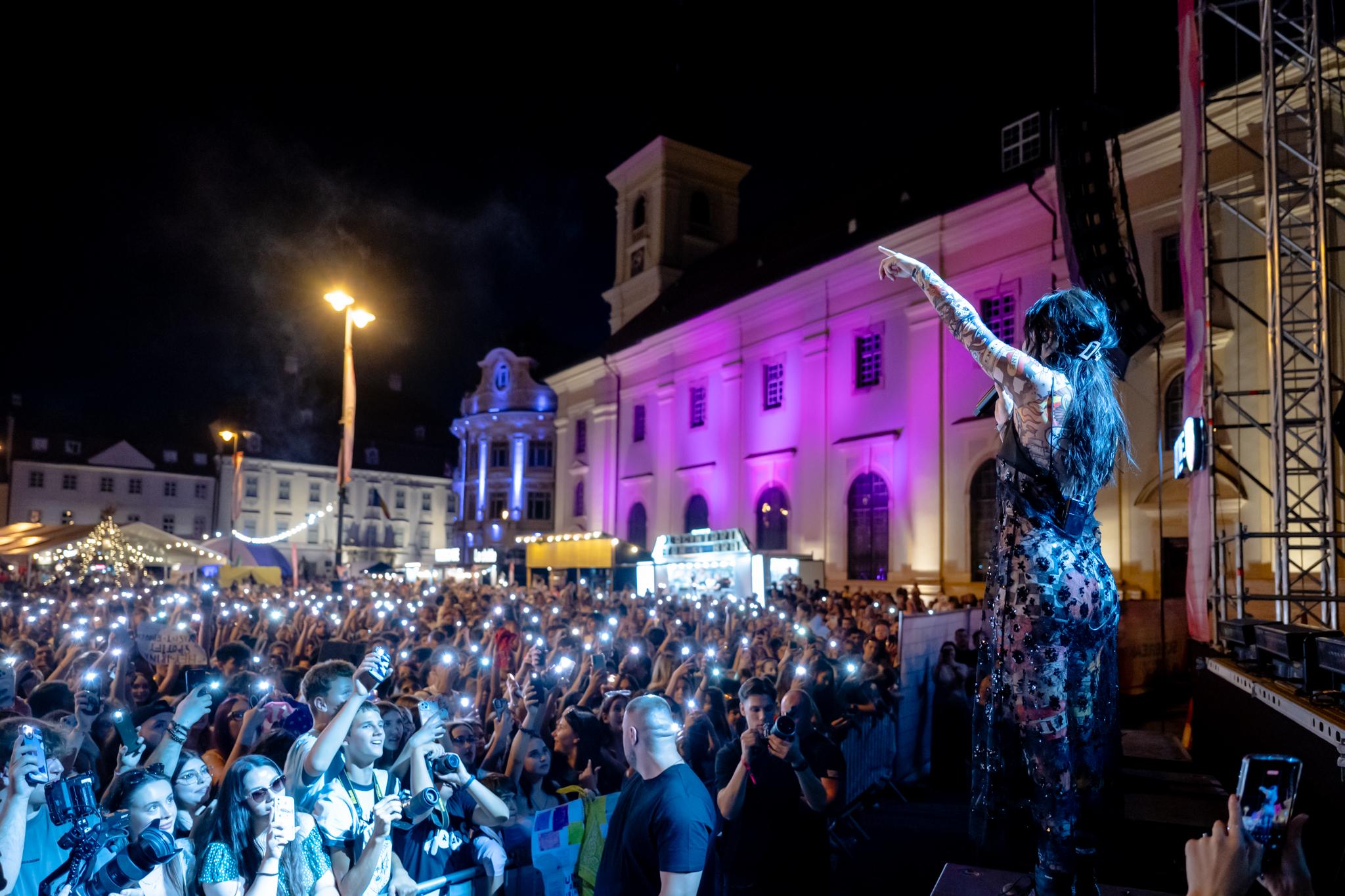 Artistă pe scenă în Piața Mare din Sibiu, arătând spre publicul cu lumini de telefon aprinse