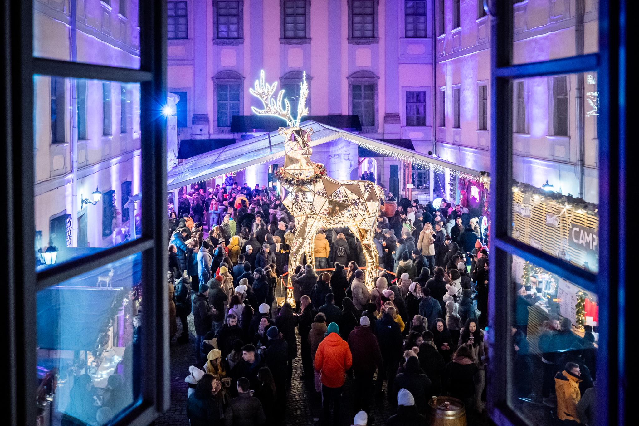 Curtea plină de oameni și renul luminos, văzute printr-o fereastră deschisă de la etaj