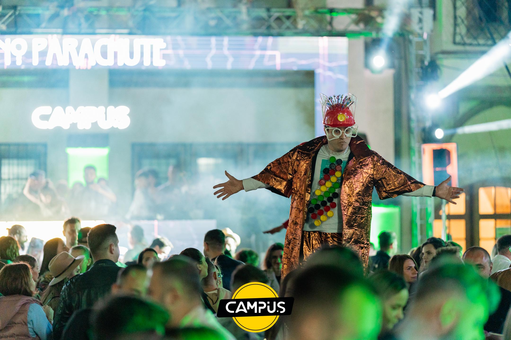Artist pe picioroange, în costum arămiu, cu brațele deschise deasupra publicului, în fața scenei